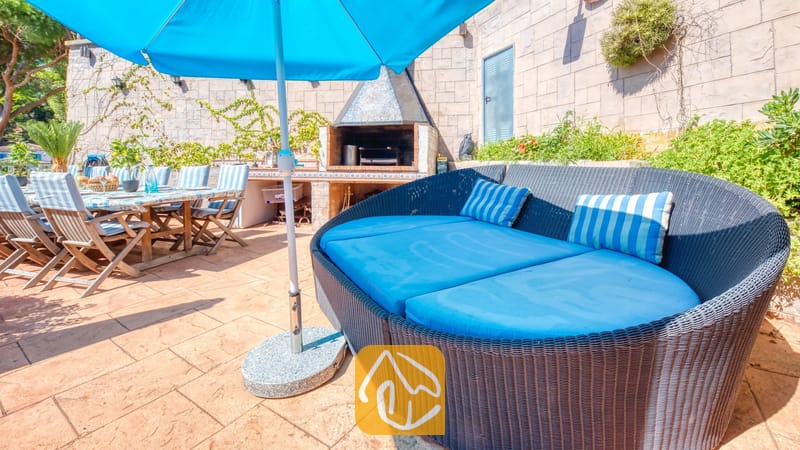 Villas de vacances Costa Brava Espagne - Villa Infinity - Canapé d'extérieur incurvé en rotin doté de coussins d'un bleu éclatant et de coussins rayés sur la terrasse carrelée de la Villa Infinity, avec un barbecue en briques et une table à manger en arrière-plan.