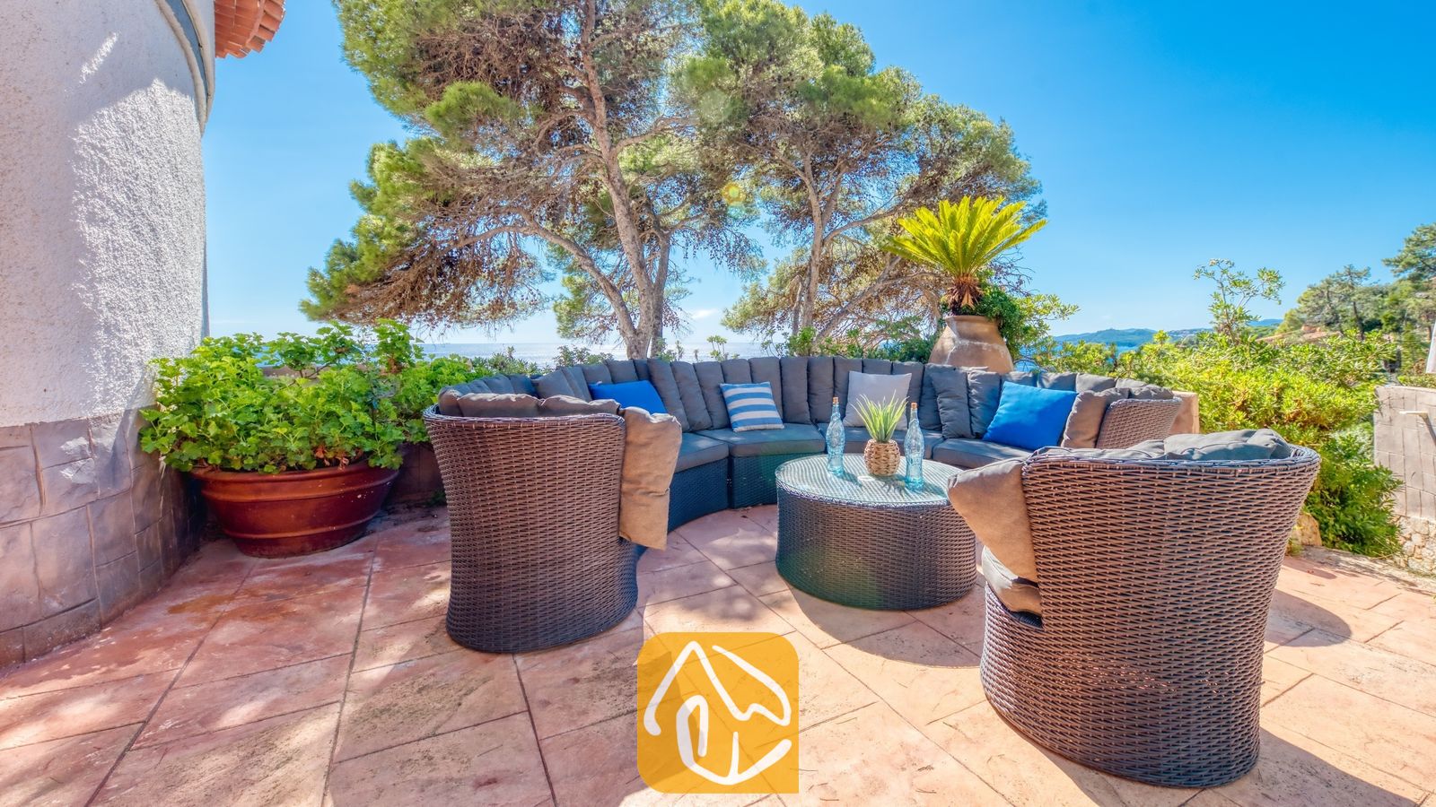 Vakantievilla's Costa Brava Spanje - Villa Infinity - Terras met zitgedeelte bij Villa Infinity, voorzien van donkere rotan fauteuils, een ronde bank met blauwe kussens en een salontafel met glazen blad onder dennenbomen.