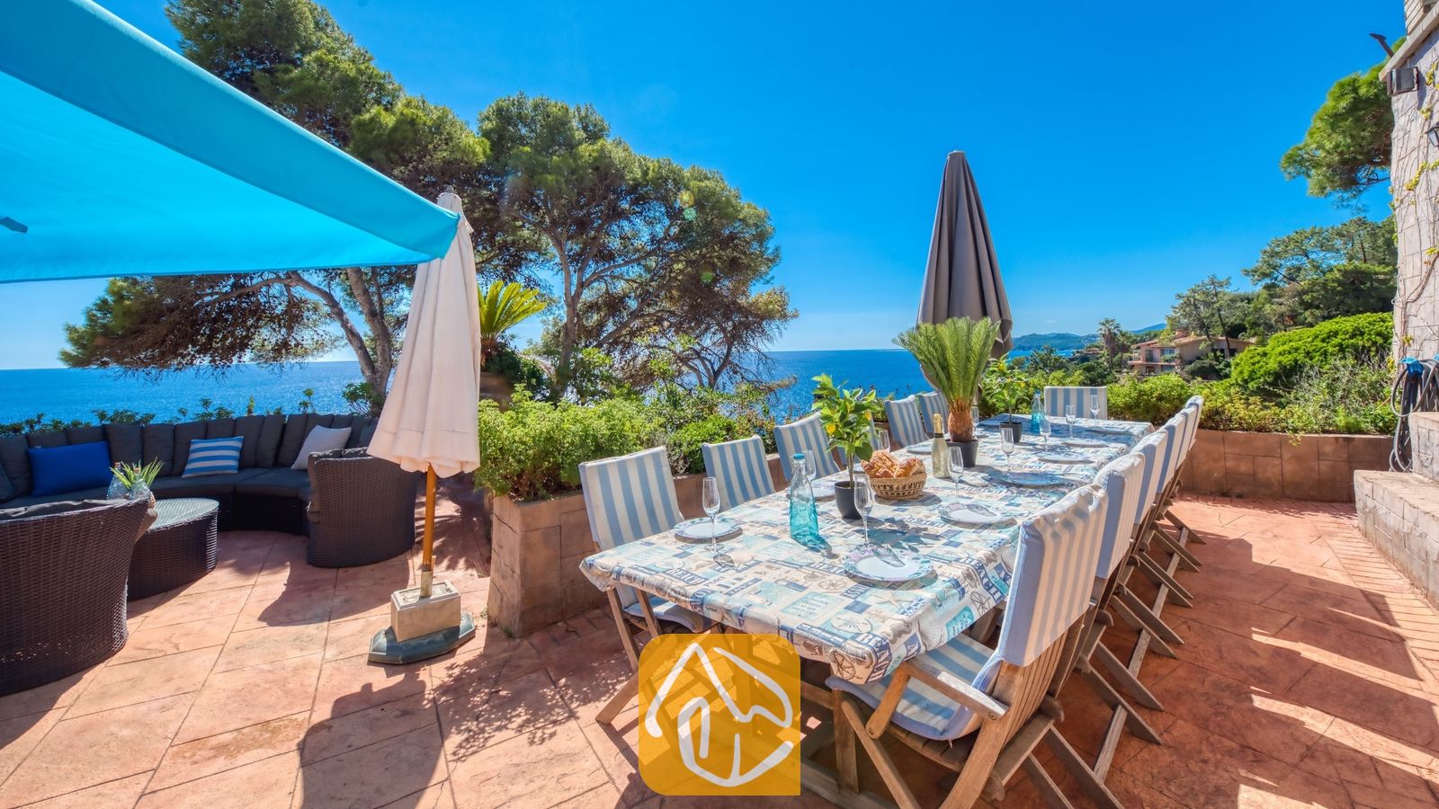 Vakantievilla's Costa Brava Spanje - Villa Infinity - Terras van Villa Infinity met een lange houten eettafel gedekt voor de maaltijd, omringd door stoelen met blauw-witte gestreepte kussens en uitzicht op zee.