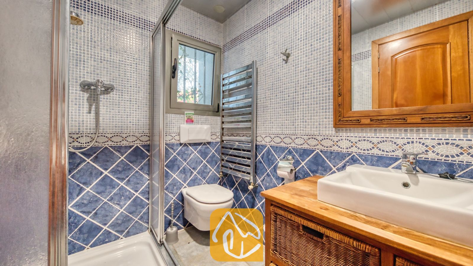 Vakantievilla's Costa Brava Spanje - Villa Infinity - Badkamer met muren van blauwe mozaïektegels, een witte keramische wastafel op een houten badkamermeubel met rieten opbergmanden, een glazen douchecabine en een hangend toilet.
