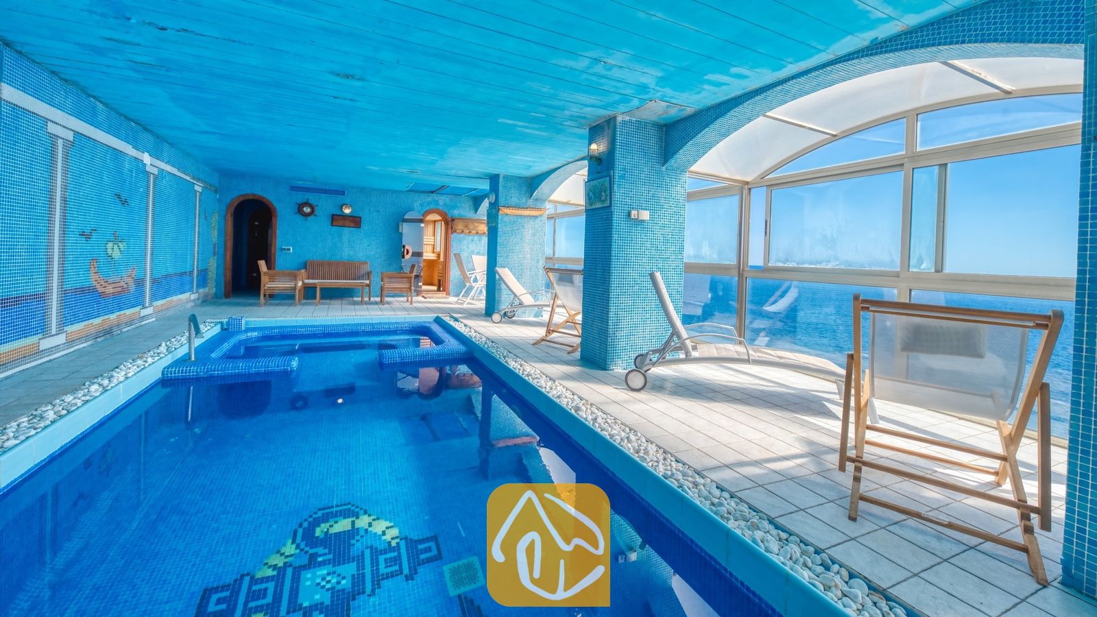 Vakantievilla's Costa Brava Spanje - Villa Infinity - Binnen zwembad met tegels bij Villa Infinity, voorzien van blauwe mozaïekmuren, witte ligstoelen en grote glazen ramen met uitzicht op zee.