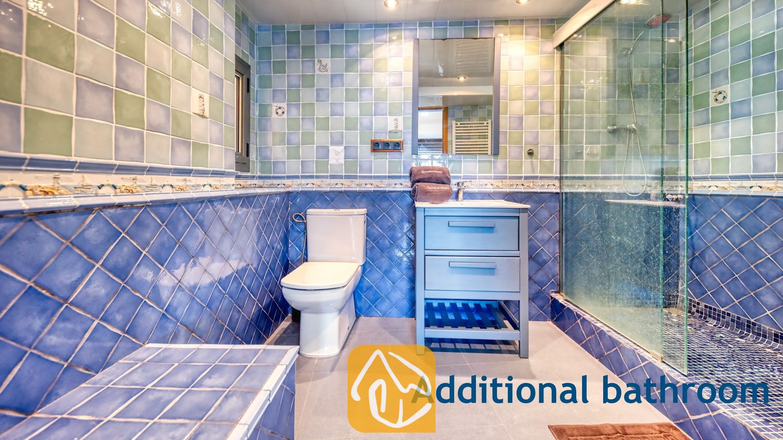 Vakantievilla's Costa Brava Spanje - Villa Infinity - Badkamer met blauwe en groene vierkante tegels, een wit toilet, een blauw badkamermeubel met wastafel en spiegel, en een glazen inloopdouche.