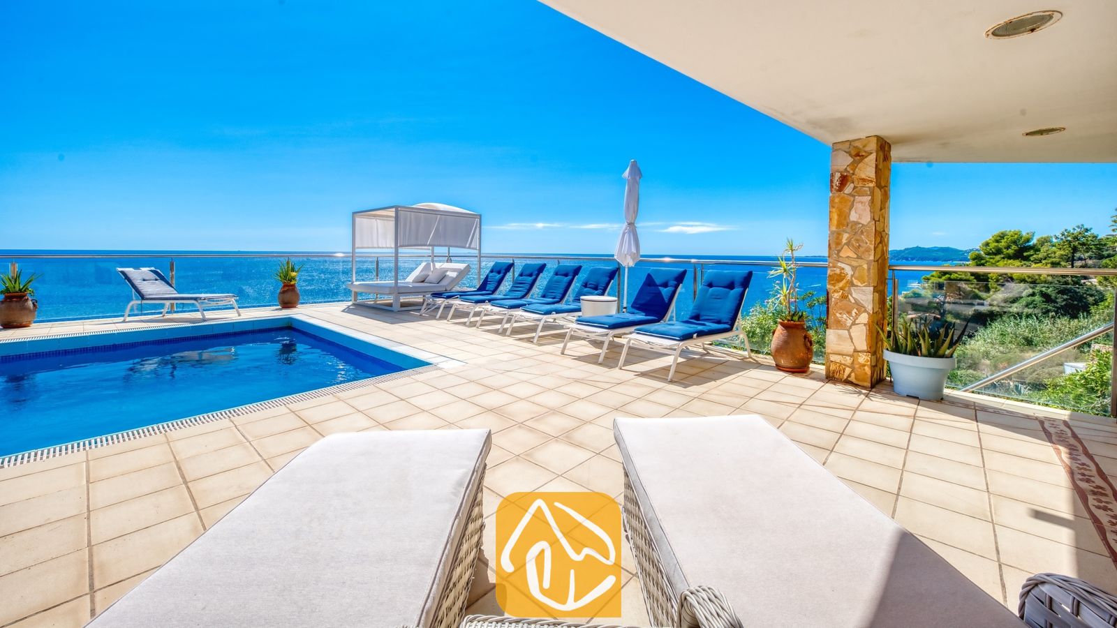 Vakantievilla's Costa Brava Spanje - Villa Infinity - Zonnig terras bij Villa Infinity met een zwembad, meerdere blauwe ligstoelen, een wit overdekt ligbed en uitzicht op zee onder een helderblauwe hemel.
