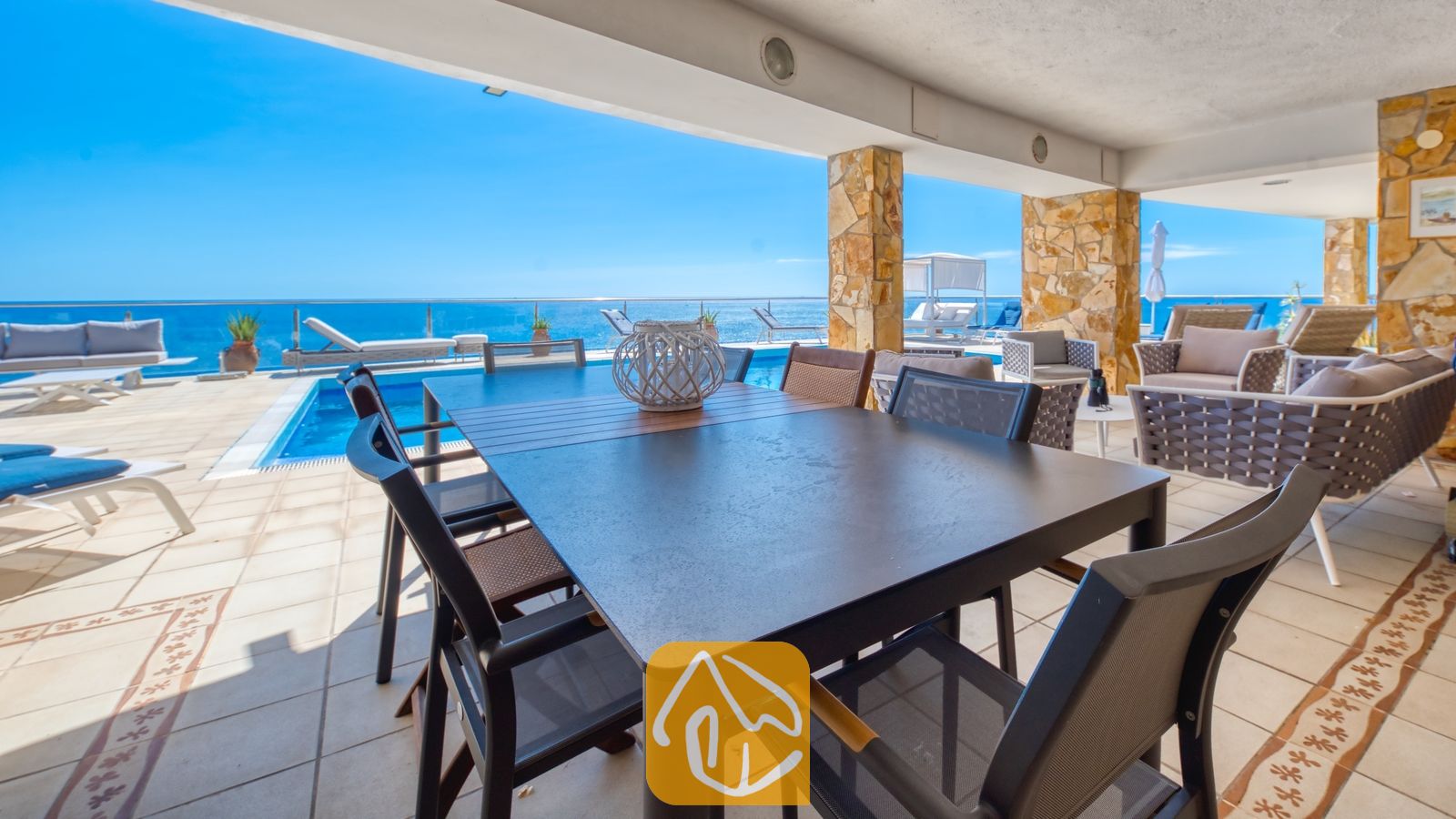 Vakantievilla's Costa Brava Spanje - Villa Infinity - Overdekt terras bij Villa Infinity met een donkere eettafel en gaasstoelen, stenen pilaren en een aangrenzend buitenzwembad met uitzicht op zee.