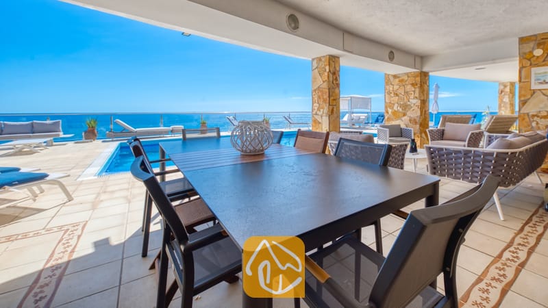 Vakantievilla's Costa Brava Spanje - Villa Infinity - Overdekt terras bij Villa Infinity met een donkere eettafel en gaasstoelen, stenen pilaren en een aangrenzend buitenzwembad met uitzicht op zee.