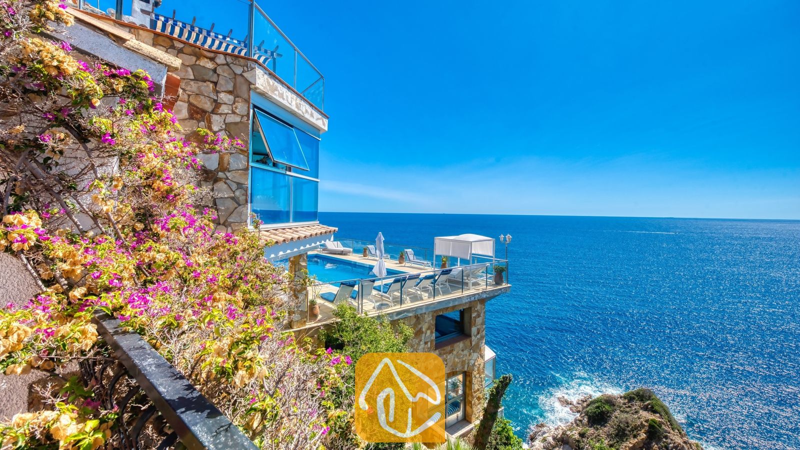 Vakantievilla's Costa Brava Spanje - Villa Infinity - Buitenkant van Villa Infinity met een stenen terras over meerdere niveaus, een zwembad, ligstoelen en zeezicht omlijst door roze bougainvillea.