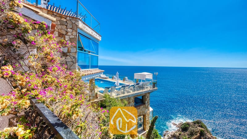 Vakantievilla's Costa Brava Spanje - Villa Infinity - Buitenkant van Villa Infinity met een stenen terras over meerdere niveaus, een zwembad, ligstoelen en zeezicht omlijst door roze bougainvillea.
