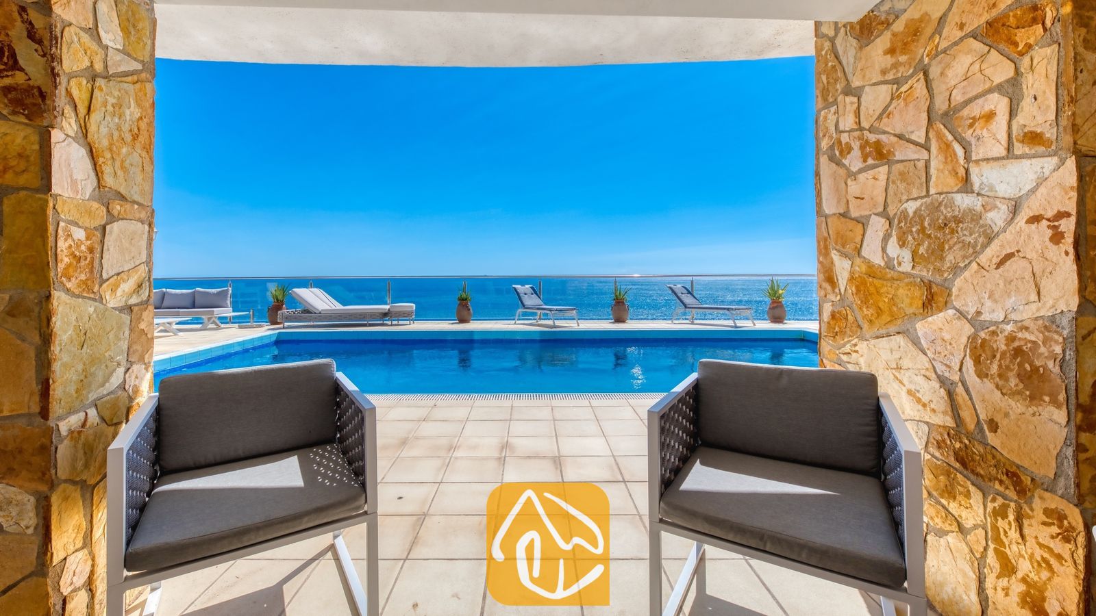 Vakantievilla's Costa Brava Spanje - Villa Infinity - Terras met uitzicht op een privézwembad en de zee door stenen pilaren bij Villa Infinity, voorzien van grijs gepositioneerde fauteuils en ligstoelen.