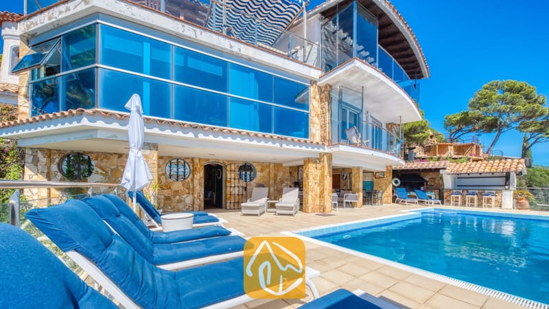 Vakantievilla's Costa Brava Spanje - Villa Infinity - Buitenaanzicht van Villa Infinity met een zwembad, blauwe ligstoelen op het stenen terras, stenen pilaren en een gebouw met meerdere verdiepingen en een glazen gevel onder een helderblauwe hemel.