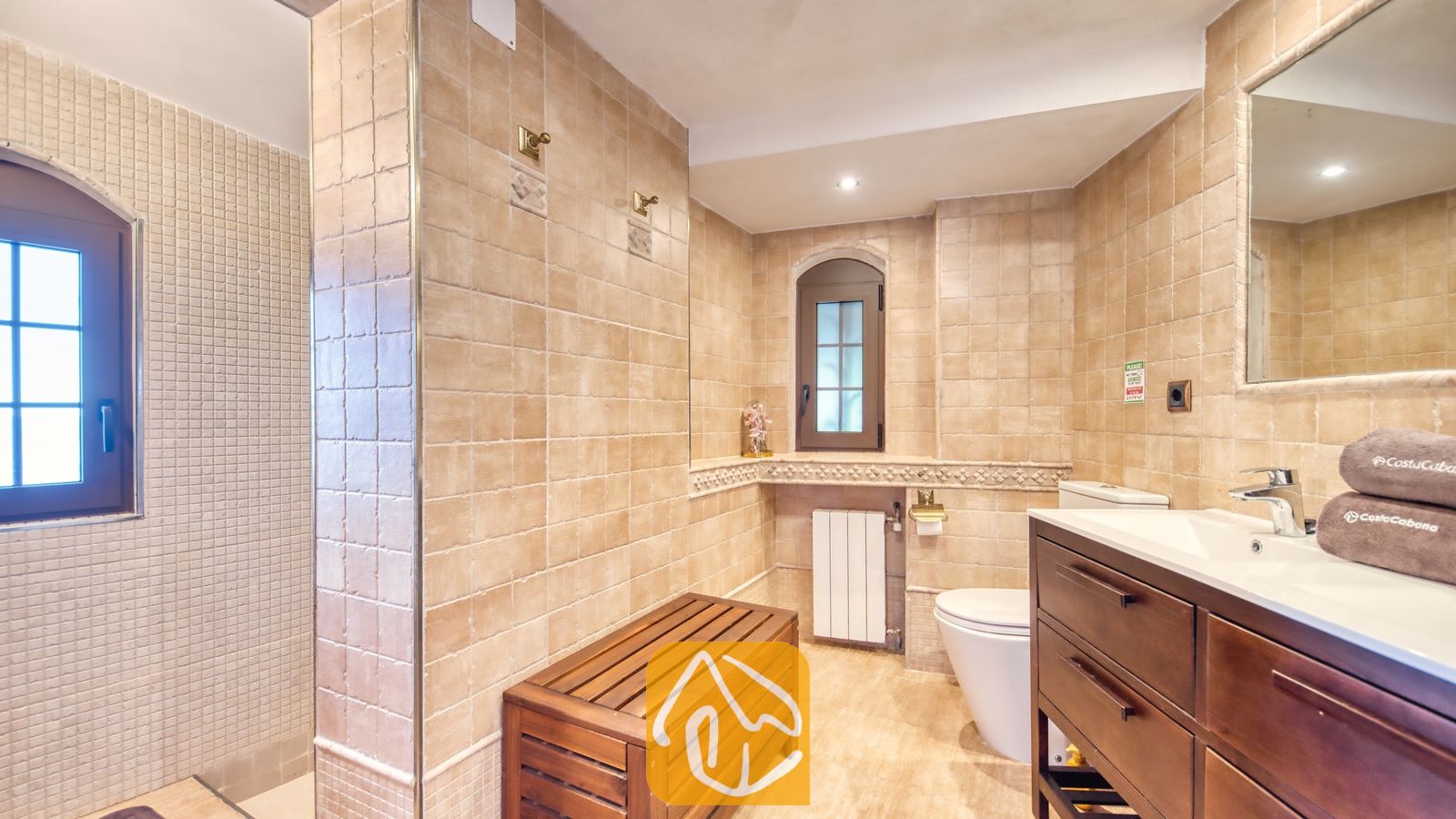 Vakantievilla's Costa Brava Spanje - Villa Infinity - Badkamer voorzien van beige vierkante keramische tegels, een houten lattenbank, een wastafelmeubel van donker hout met een witte wastafel en een spiegel aan de muur.