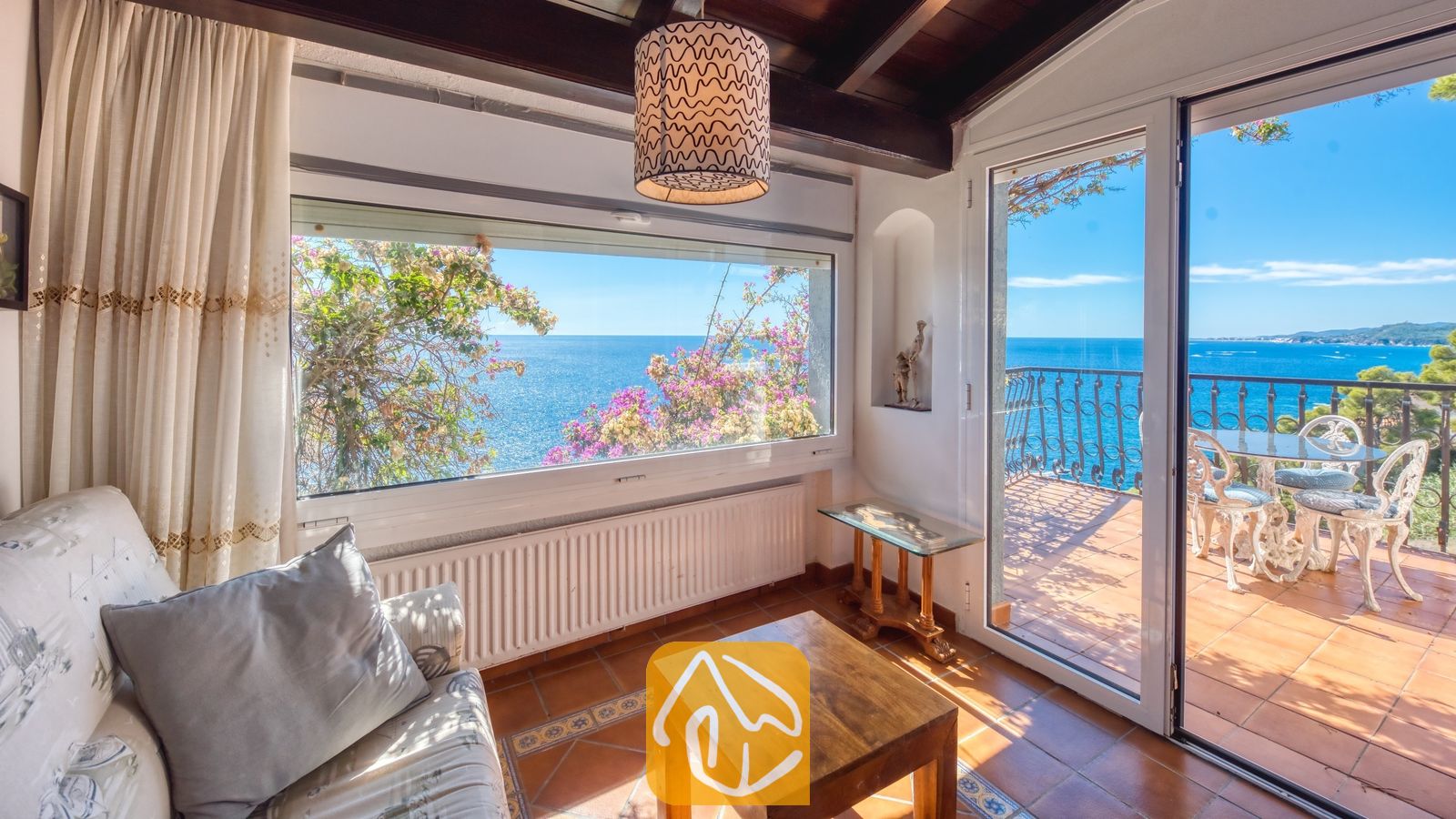 Vakantievilla's Costa Brava Spanje - Villa Infinity - Woonkamer met uitzicht op zee, een houten salontafel, een witte bank en een glazen schuifpui die uitkomt op een balkon met witte metalen stoelen.