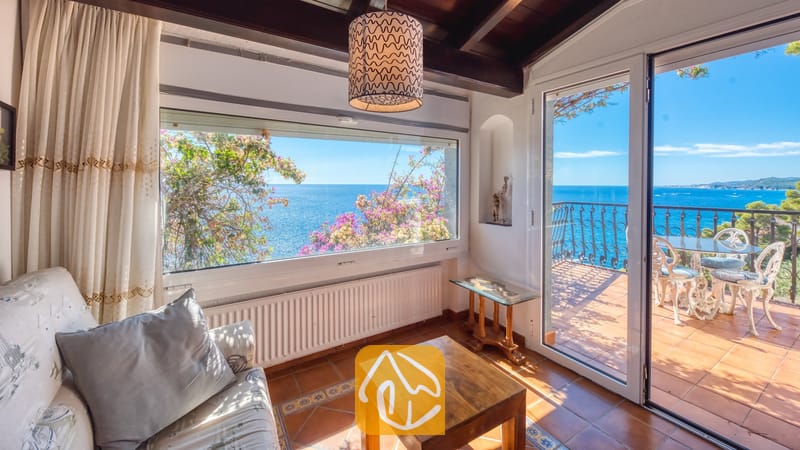 Villas de vacances Costa Brava Espagne - Villa Infinity - Salon avec vue sur mer, table basse en bois, canapé blanc et porte-fenêtre coulissante s'ouvrant sur un balcon doté de chaises en métal blanc.