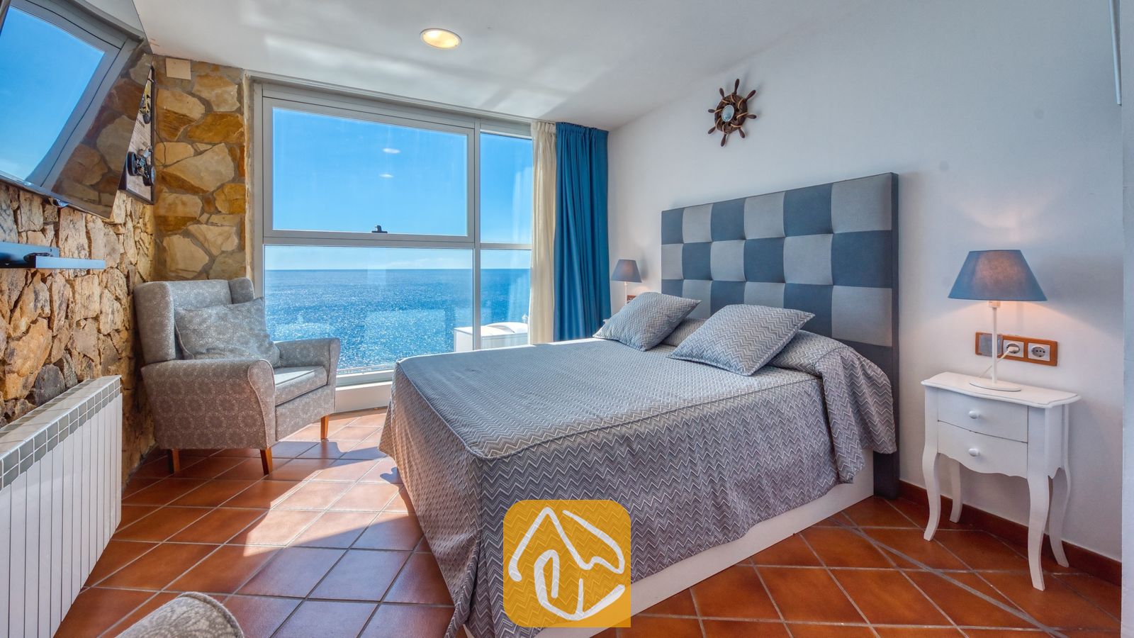 Vakantievilla's Costa Brava Spanje - Villa Infinity - Slaapkamer met een groot bed, een grijs geruit hoofdeinde, een fauteuil, een stenen accentmuur en een groot raam met uitzicht op zee.