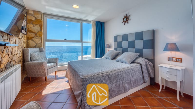 Villas de vacances Costa Brava Espagne - Villa Infinity - Chambre avec un grand lit, une tête de lit à motifs carreaux gris, un fauteuil, un mur d'accent en pierre et une grande fenêtre offrant une vue sur la mer.