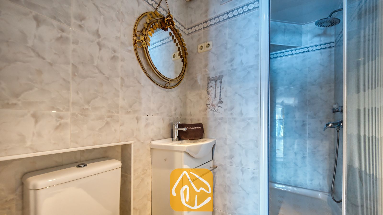 Vakantievilla's Costa Brava Spanje - Villa Infinity - Betegelde badkamer met een kleine witte wasbak, een decoratieve wandspiegel met een gouden lijst en een glazen doucheruimte met een stortdouche.