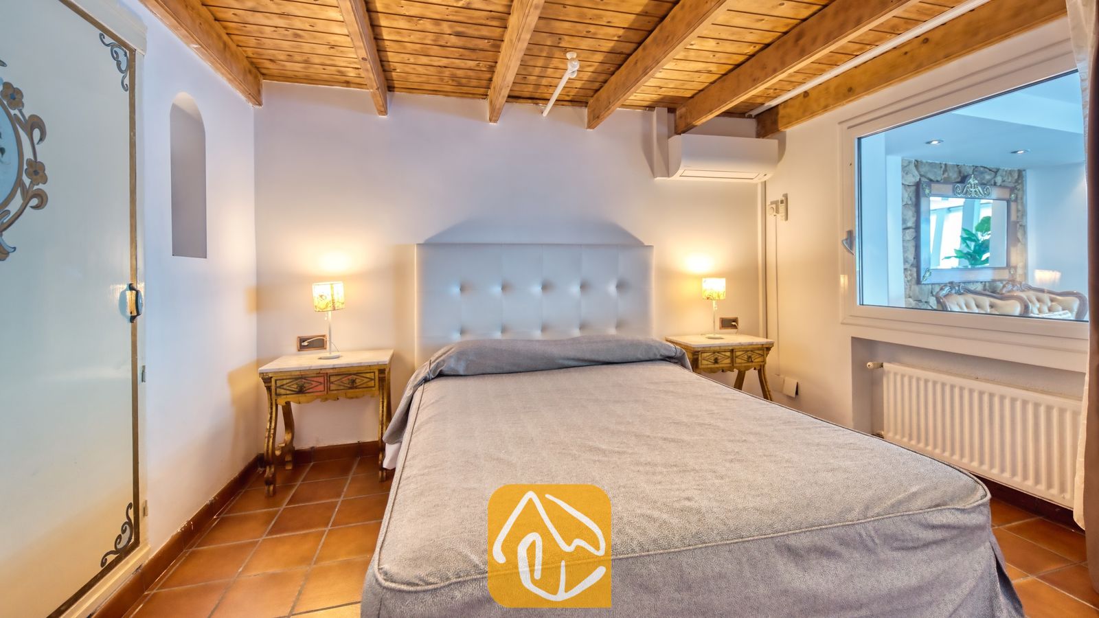 Vakantievilla's Costa Brava Spanje - Villa Infinity - Slaapkamer met een wit getuft hoofdeinde, een grijs sprei, houten plafondbalken en tegelvloeren.