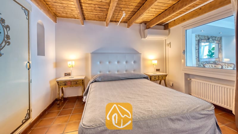 Villas de vacances Costa Brava Espagne - Villa Infinity - Chambre à coucher avec une tête de lit capitonnée blanche, un couvre-lit gris, des poutres apparentes en bois et un sol en carrelage.