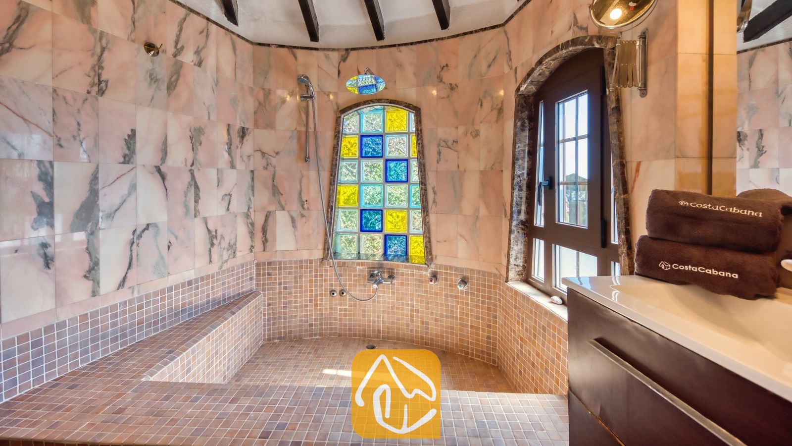 Vakantievilla's Costa Brava Spanje - Villa Infinity - Ruime inloopdouche met roze marmeren muren, bruine mozaïekvloertegels en een kleurrijk glas-in-loodraam boven chromen armaturen.