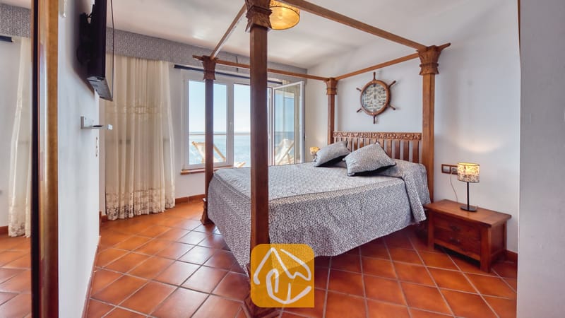 Villas de vacances Costa Brava Espagne - Villa Infinity - Chambre avec lit baldaquin en bois, parure de lit à motifs, sols carrelés et fenêtre avec vue sur la mer.