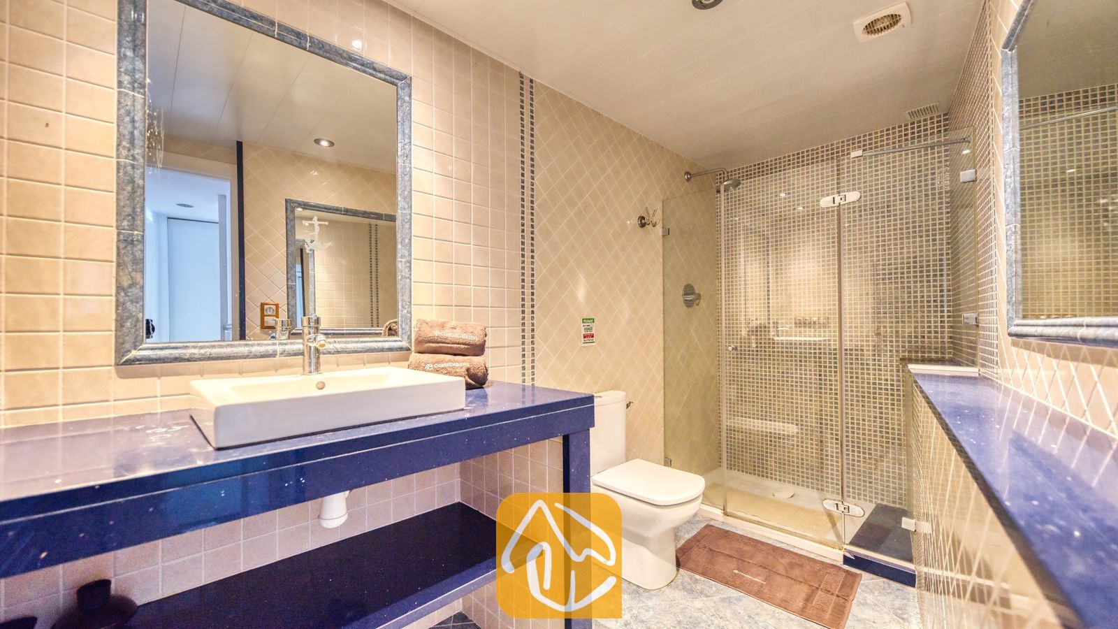 Vakantievilla's Costa Brava Spanje - Villa Infinity - Badkamer met een witte opzetwastafel op een blauw badkamermeubel, een grote wandspiegel, een toilet en een glazen inloopdouche met mozaïektegels.