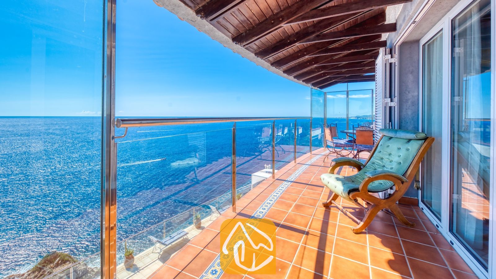 Vakantievilla's Costa Brava Spanje - Villa Infinity - Overdekt terras bij Villa Infinity met een groene houten fauteuil, tegelvloer, glazen balustrade en vrij uitzicht op zee onder een helderblauwe hemel.