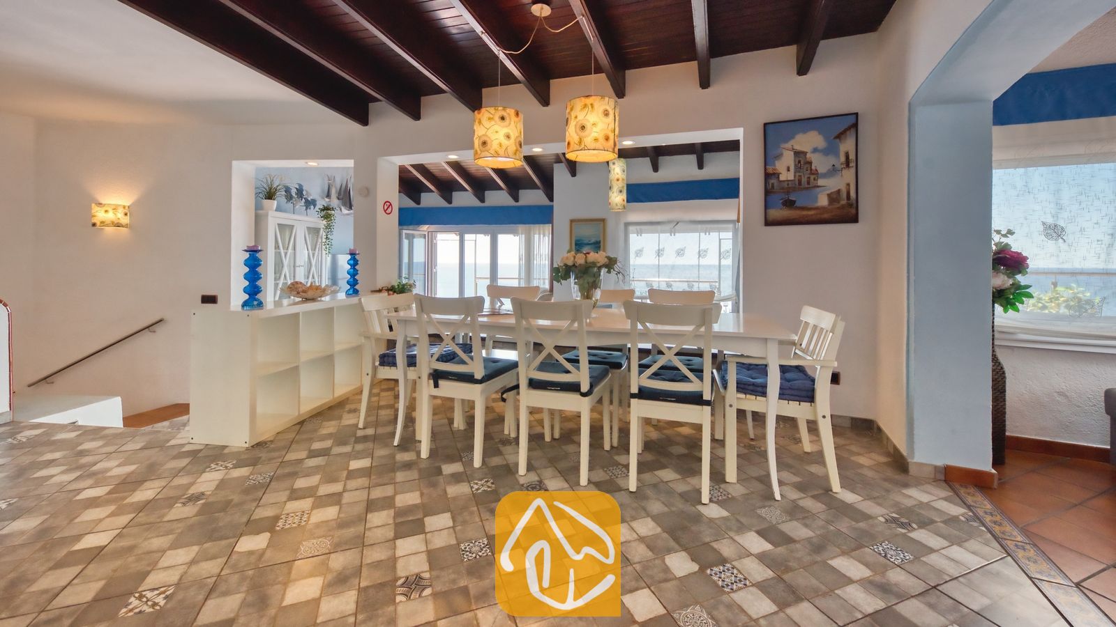 Vakantievilla's Costa Brava Spanje - Villa Infinity - Eetgedeelte met een lange witte tafel en bijpassende stoelen, donkere houten balkenplafonds en vloeren met patroon tegels in Villa Infinity.