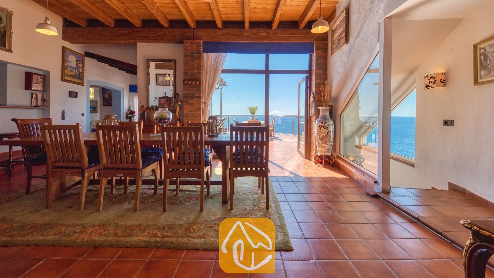 Vakantievilla's Costa Brava Spanje - Villa Infinity - Houten eettafel en stoelen op een gedessineerd vloerkleed in Villa Infinity, voorzien van zichtbare houten plafondbalken en grote ramen die uitzicht bieden op zee.