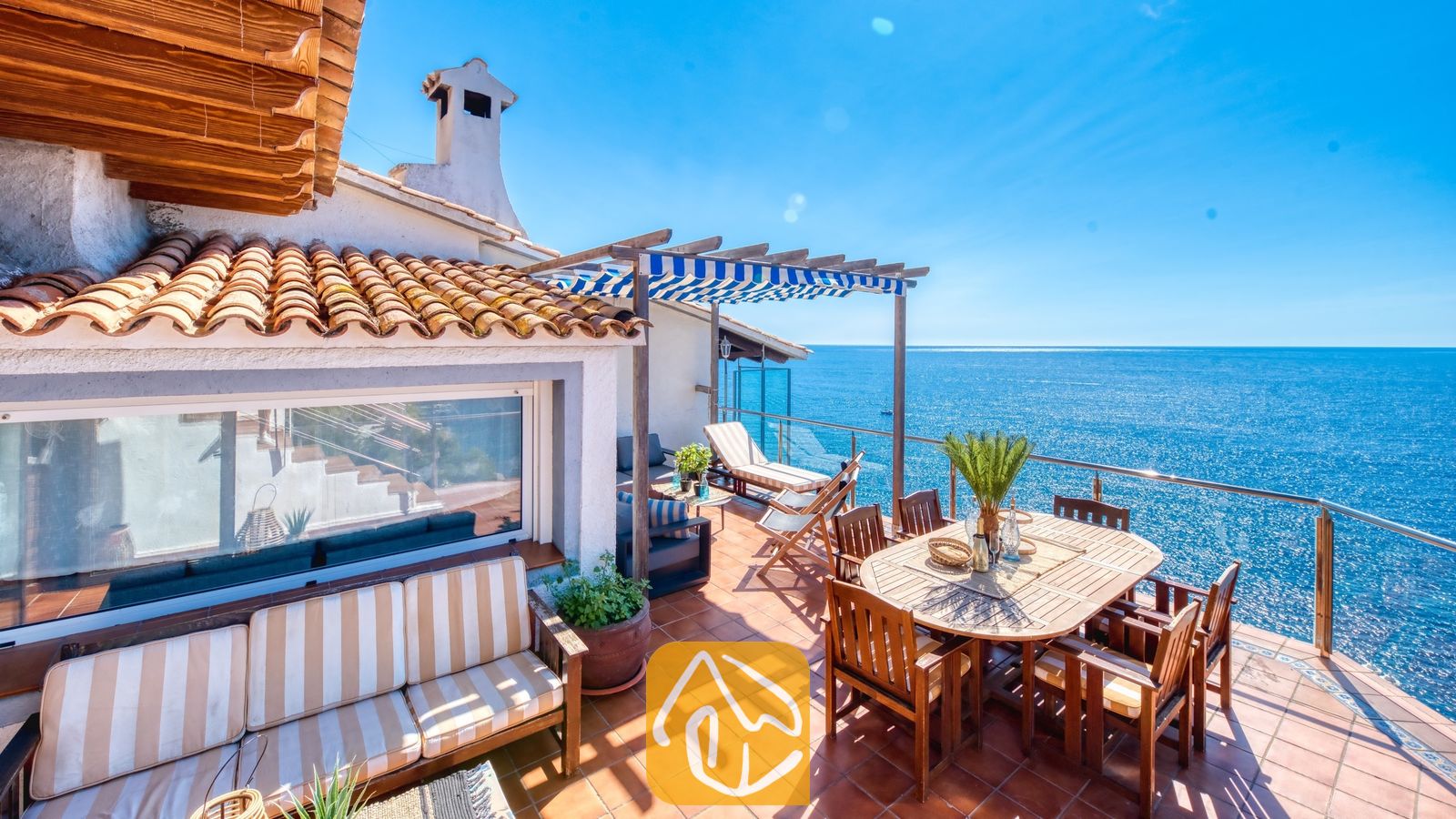 Vakantievilla's Costa Brava Spanje - Villa Infinity - Zonnig buitenterras bij Villa Infinity, voorzien van een houten eettafel met stoelen, terracotta tegelvloer en een panoramisch uitzicht op zee onder een strakke, blauwe hemel.