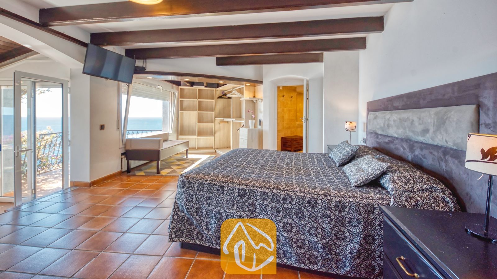 Vakantievilla's Costa Brava Spanje - Villa Infinity - Slaapkamer met donker beddengoed met motief, zichtbare houten balken aan het plafond en terracotta plavuizen in Villa Infinity