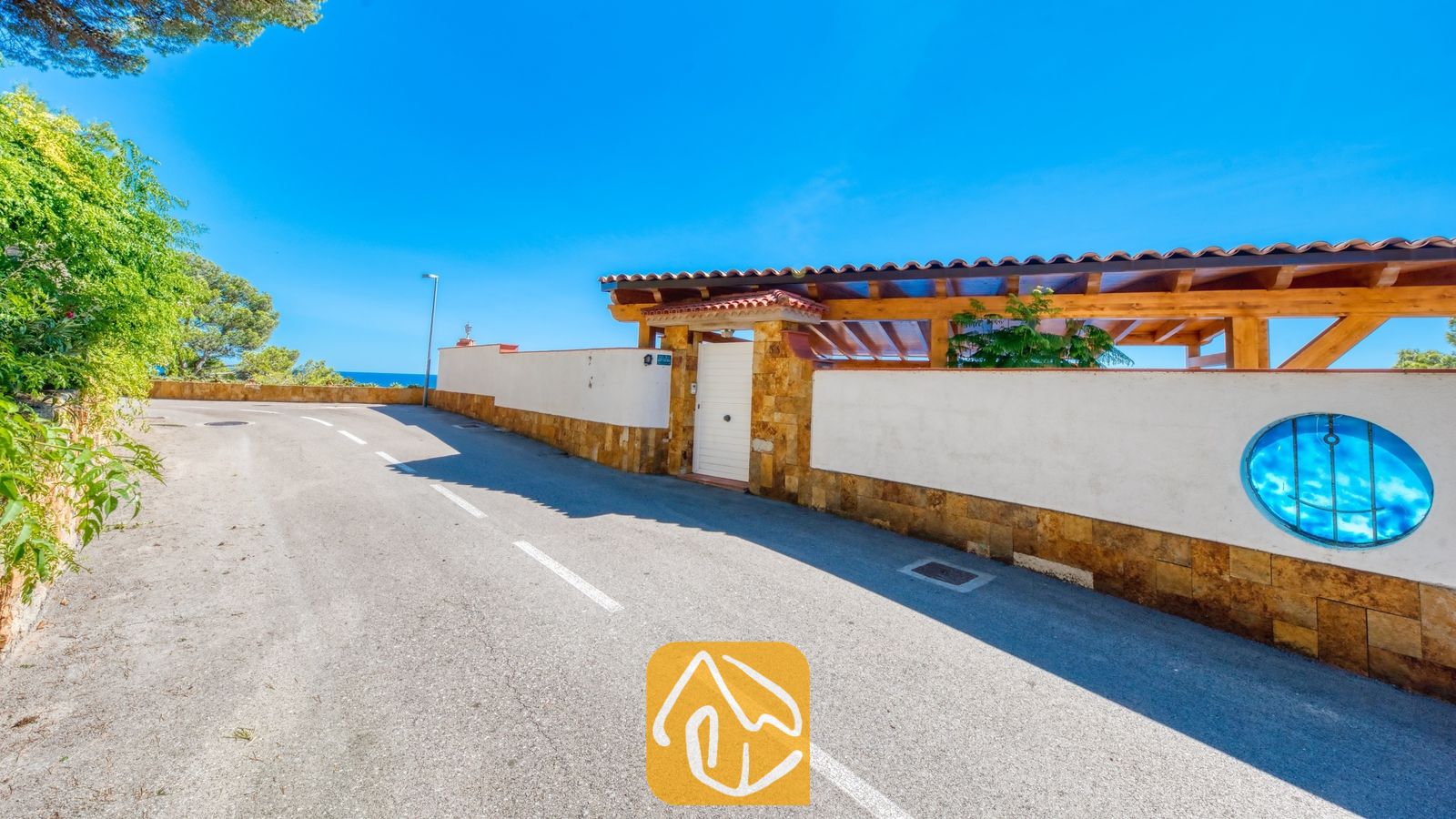 Vakantievilla's Costa Brava Spanje - Villa Infinity - Buiteningang van Villa Infinity met een geplaveide straat langs een witte erfafscheiding met stenen accenten, een houten carportdak en een rond blauw siervenster.