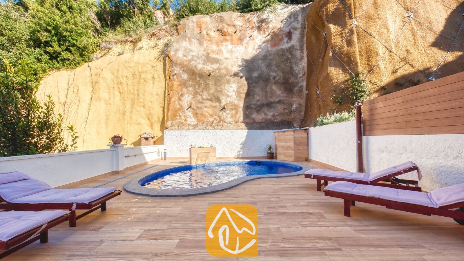 Vakantievilla's Costa Brava Spanje - Villa Blanca - Privézwembad bij Villa Blanca met blauwe mozaïektegels, gelegen op een betegeld terras omlijst door een hoge rotswand en voorzien van houten ligstoelen met paarse kussens.