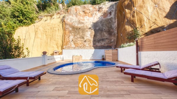 Vakantievilla's Costa Brava Spanje - Villa Blanca - Privézwembad bij Villa Blanca met blauwe mozaïektegels, gelegen op een betegeld terras omlijst door een hoge rotswand en voorzien van houten ligstoelen met paarse kussens.