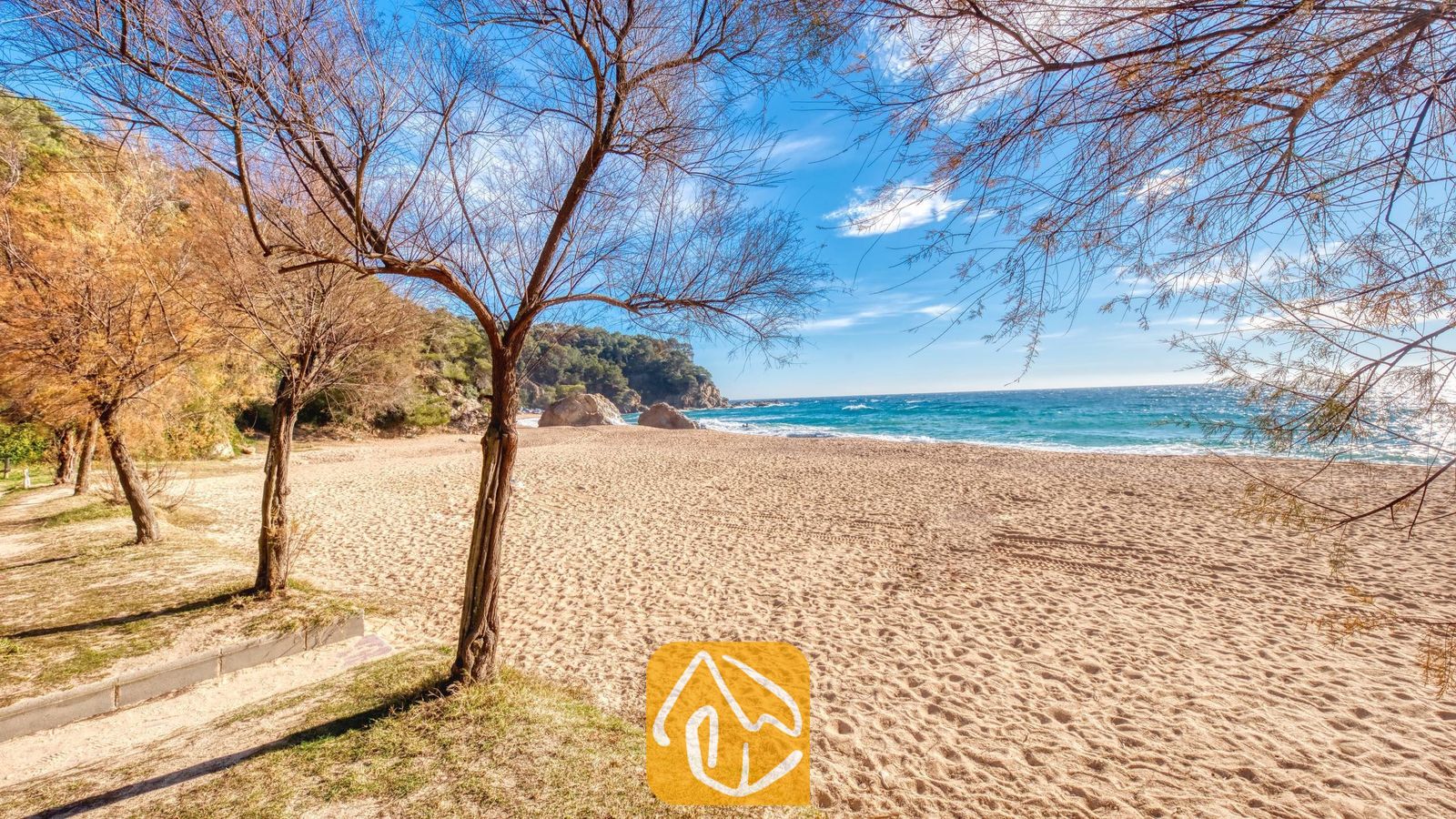 Vakantievilla's Costa Brava Spanje - Villa Blanca - Zandstrand met kale bomen die de blauwe zee en de rotsachtige kustlijn omlijsten nabij Villa Blanca in Spanje
