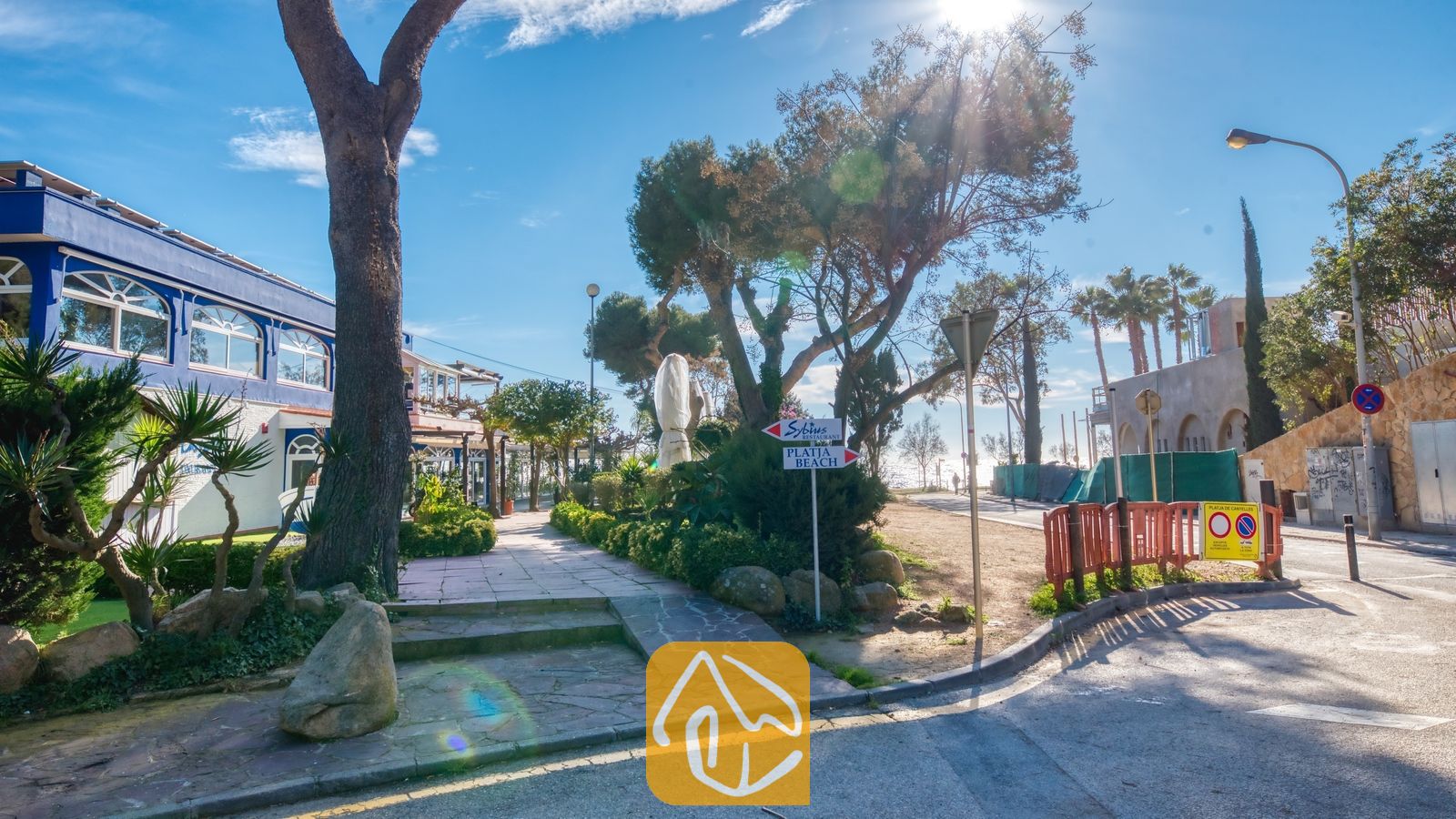 Vakantievilla's Costa Brava Spanje - Villa Blanca - Zonnige kuststraat vlak bij Villa Blanca, voorzien van een verhard wandelpad, volwassen bomen, bewegwijzering naar het strand en blauw-witte gebouwen onder een heldere hemel.