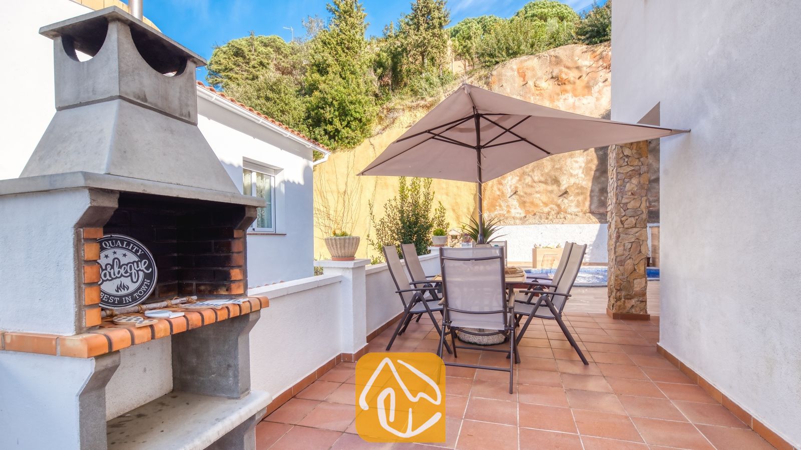 Vakantievilla's Costa Brava Spanje - Villa Blanca - Buitenbesterras van Villa Blanca met een stenen barbecue, een tuintafel met zes stoelen en een grote parasol op een tegelvloer.