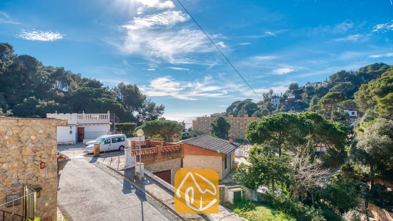 Vakantievilla's Costa Brava Spanje - Villa Blanca - Buitenaanzicht van Villa Blanca met een verharde oprijlaan, geparkeerde voertuigen, stenen muren en omringende groene heuvels onder een blauwe lucht met verspreide wolken.