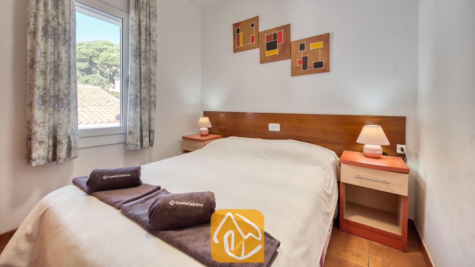 Vakantievilla's Costa Brava Spanje - Villa Blanca - Slaapkamer met een tweepersoonsbed voorzien van wit beddengoed, opgerolde bruine handdoeken, houten nachtkastjes met lampen en drie abstracte kunstwerken aan de muur.