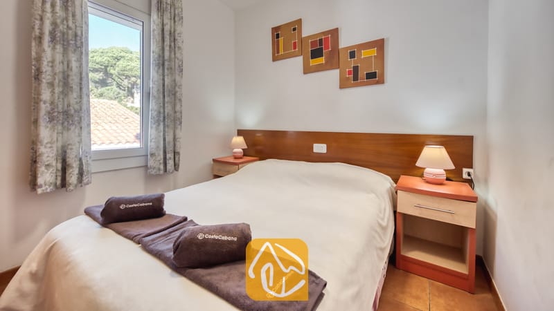 Vakantievilla's Costa Brava Spanje - Villa Blanca - Slaapkamer met een tweepersoonsbed voorzien van wit beddengoed, opgerolde bruine handdoeken, houten nachtkastjes met lampen en drie abstracte kunstwerken aan de muur.