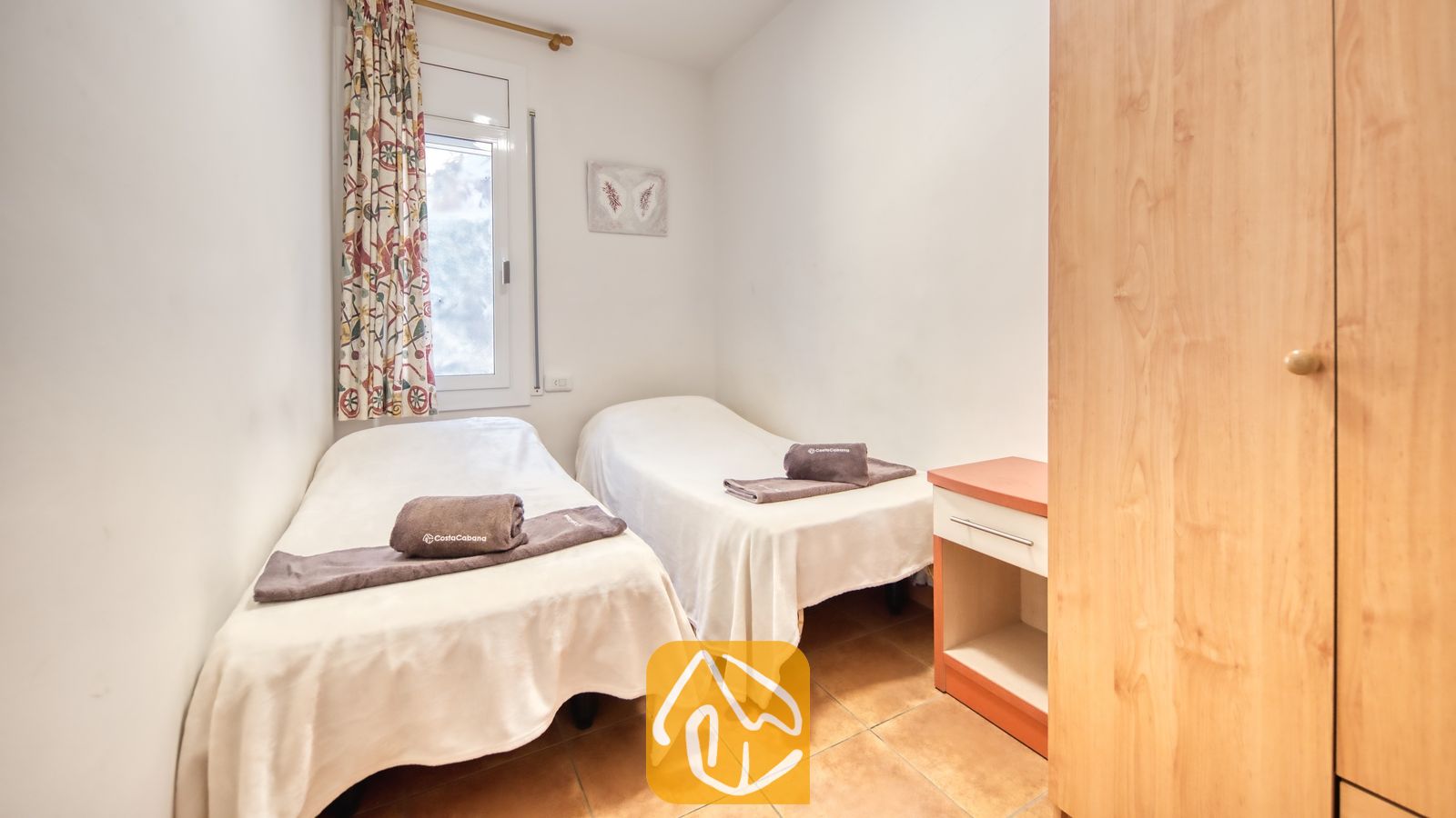 Vakantievilla's Costa Brava Spanje - Villa Blanca - Slaapkamer met twee eenpersoonsbedden opgemaakt met witte plaids, opgevouwen bruine handdoeken, gordijnen met een motief en een houten kledingkast.