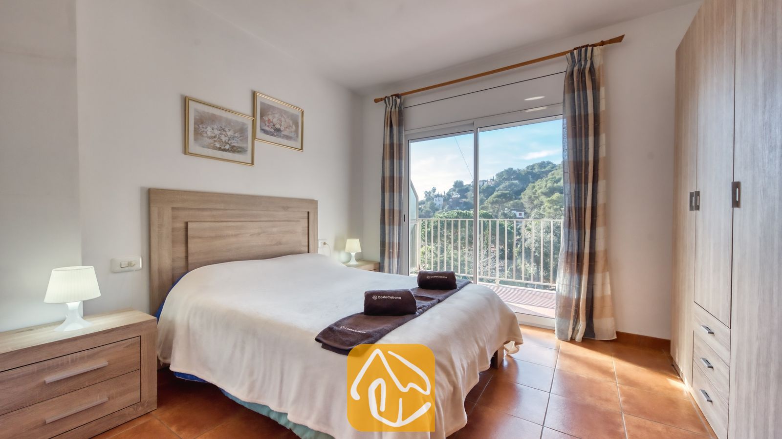 Vakantievilla's Costa Brava Spanje - Villa Blanca - Slaapkamer met een tweepersoonsbed, houten hoofdbord, twee nachtkastjes met lampen en een glazen schuifpui die uitkomt op een balkon met uitzicht op groene heuvels.