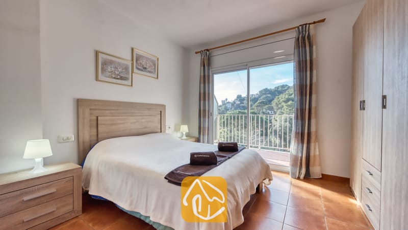 Vakantievilla's Costa Brava Spanje - Villa Blanca - Slaapkamer met een tweepersoonsbed, houten hoofdbord, twee nachtkastjes met lampen en een glazen schuifpui die uitkomt op een balkon met uitzicht op groene heuvels.