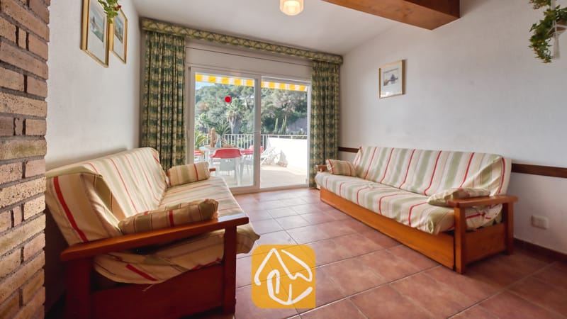 Vakantievilla's Costa Brava Spanje - Villa Blanca - Woonkamer met twee gestreepte houten banken, een tegelvloer en glazen schuifpuien die uitkomen op een terras bij Villa Blanca