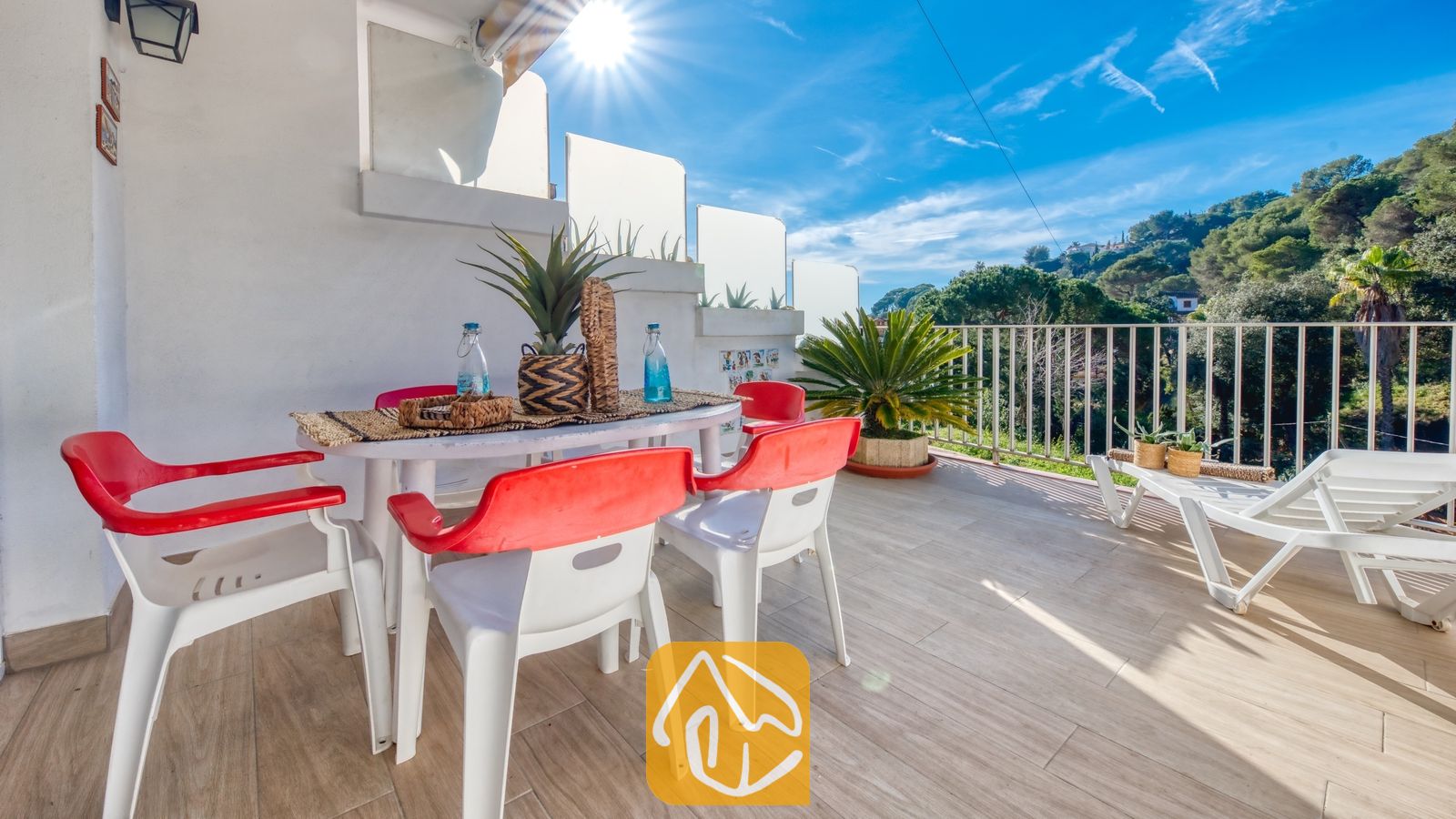 Vakantievilla's Costa Brava Spanje - Villa Blanca - Zonnig terras bij Villa Blanca met een eettafel, rood-witte kunststof stoelen en twee witte ligstoelen, met uitzicht op groene heuvels onder een blauwe lucht