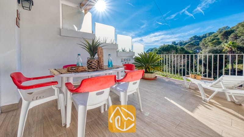Vakantievilla's Costa Brava Spanje - Villa Blanca - Zonnig terras bij Villa Blanca met een eettafel, rood-witte kunststof stoelen en twee witte ligstoelen, met uitzicht op groene heuvels onder een blauwe lucht