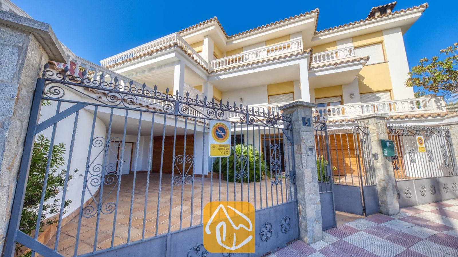 Vakantievilla's Costa Brava Spanje - Villa Baileys - De buitenzijde van Villa Baileys, met een geel-witte gebouw van meerdere verdiepingen achter een sierlijk grijs metalen toegangshek en een betegelde oprijlaan onder een helderblauwe hemel.