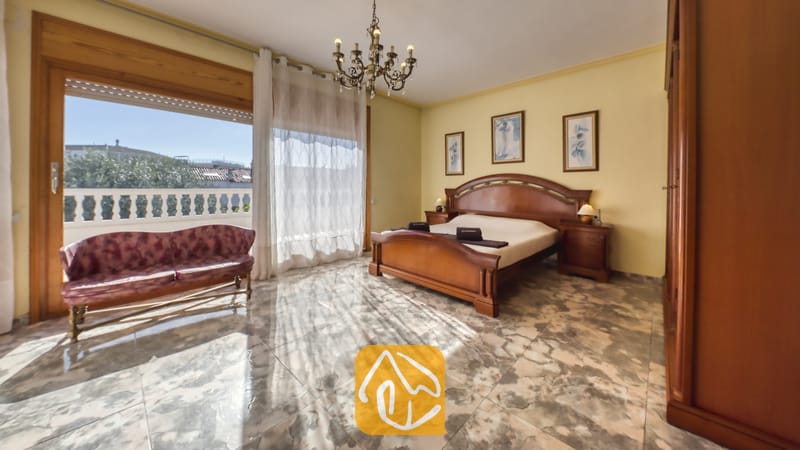 Vakantievilla's Costa Brava Spanje - Villa Baileys - Slaapkamer met een houten bedframe, geglazuurde tegels met patroon en een glazen schuifdeur die uitkomt op een zonnig balkon.