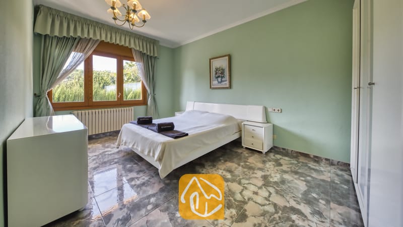Ferienhäuser Costa Brava Spanien - Villa Baileys - Schlafzimmer mit einem weißen Doppelbett, grünen Wänden, Marmorfliesenboden und einem Fenster mit grünen Vorhängen.