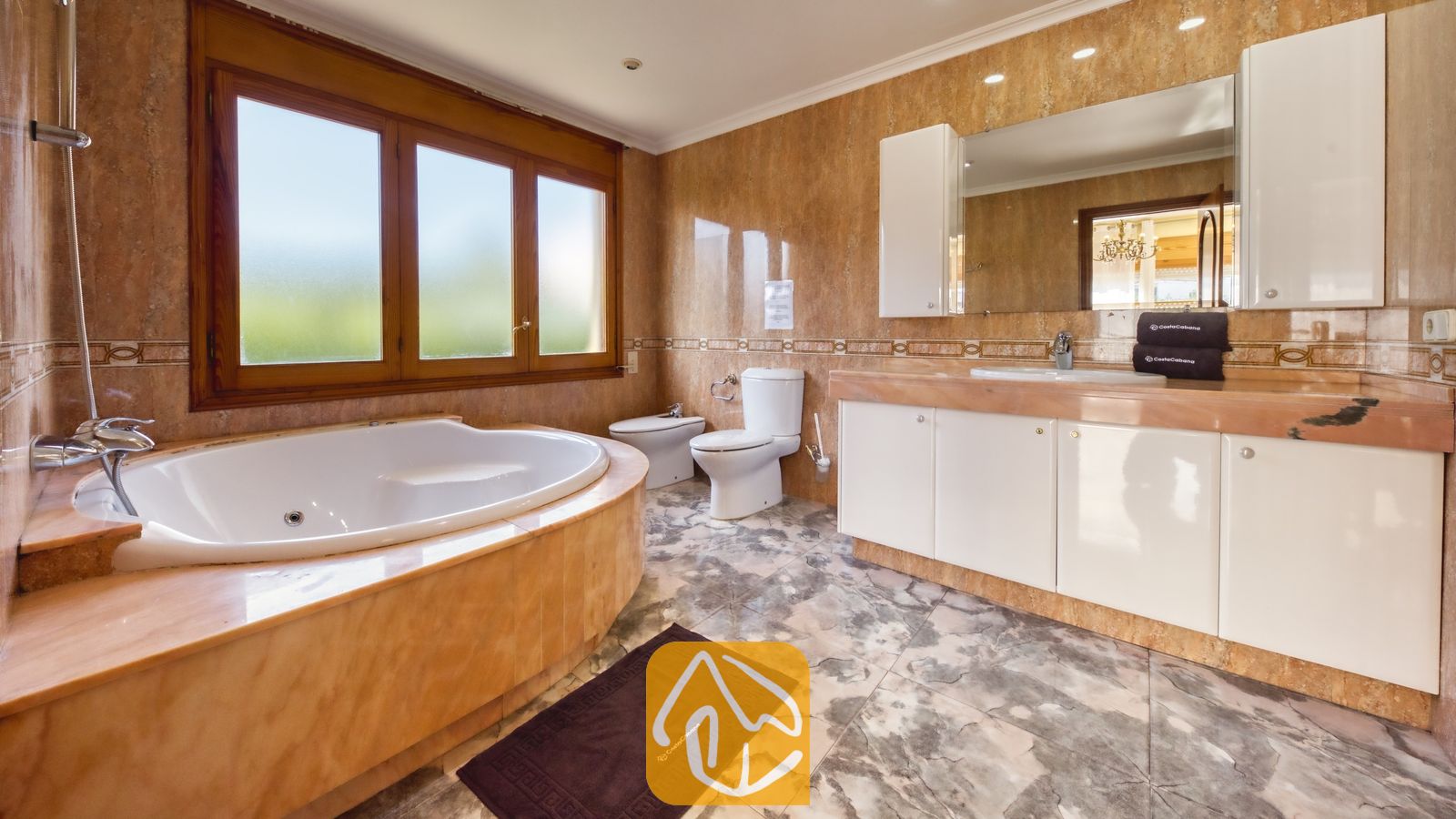 Vakantievilla's Costa Brava Spanje - Villa Baileys - Badkamer voorzien van een hoekbad, witte badkamermeubels, met marmer betegelde muren en een groot raam met een houten kozijn.