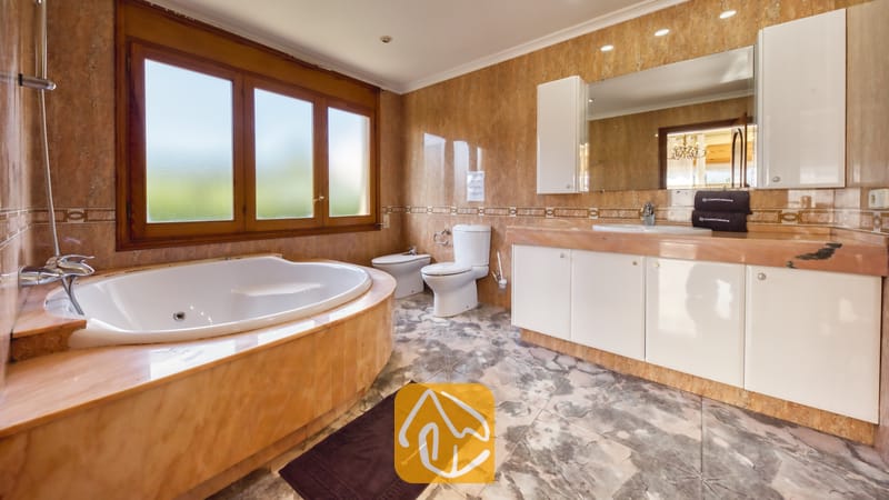 Ferienhäuser Costa Brava Spanien - Villa Baileys - Badezimmer mit einer Eckbadewanne, weißen Badmöbeln, marmorgefliesten Wänden und einem großen, holzgerahmten Fenster.