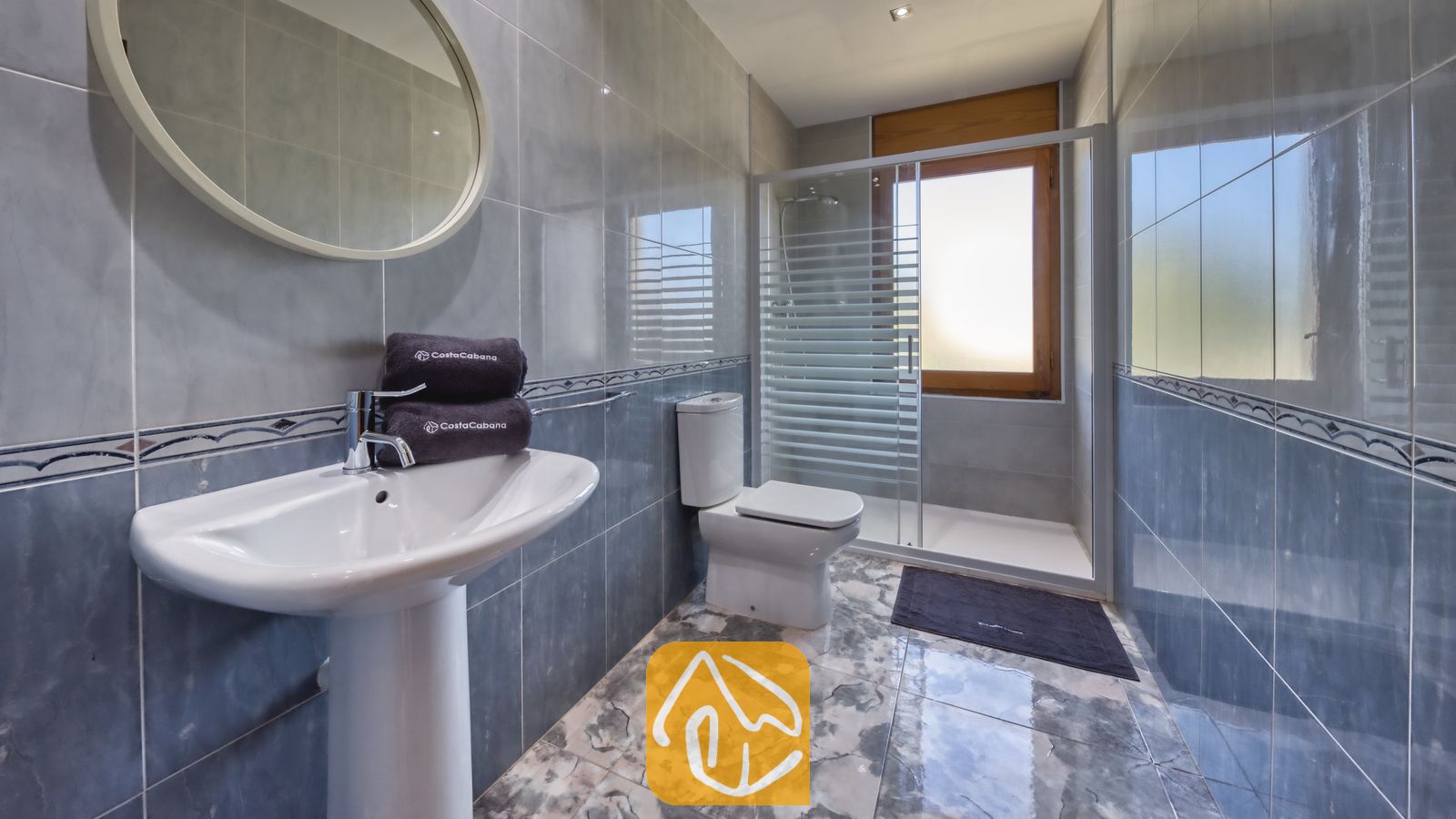 Vakantievilla's Costa Brava Spanje - Villa Baileys - Betegelde badkamer met een wastafel op zuil, een ronde spiegel, een toilet en een glazen doucheruimte met een matglas raam.